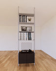 Floor-to-ceiling room divider shelving unit with lower storage cabinets with black shelves and satin vertical posts, one bay, 24-inch bay width, six open shelf levels, floor-to-ceiling supports.