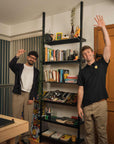 Floor-to-ceiling room divider shelving unit with black shelves and satin vertical posts, one bay, 24-inch bay width, six open shelf levels, floor-to-ceiling supports, styled in a room setting with books and decor.